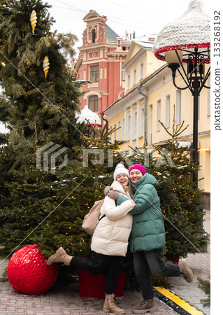 Two women in winter coats hug playfully among Christmas trees decorated with warm lights on a festive city street 133268192