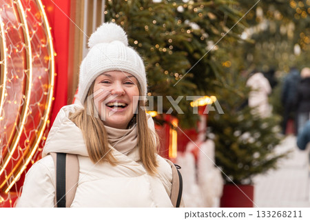 A smiling woman in a white winter coat and knitted hat stands beside festive red decorations and illuminated Christmas trees A smiling woman in a white winter coat and knitted hat stands beside festive red decorations and illuminated Christmas trees 133268211