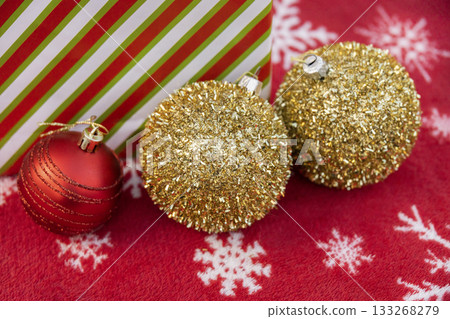 Three Christmas baubles, two gold and one red, lie beside a striped gift box on a red blanket with a snowflake pattern. The image creates a warm festive atmosphere for Christmas Three Christmas baubles, two gold and one red, lie beside a striped gift box on a red blanket with a snowflake pattern. The image creates a warm festive atmosphere for Christmas 133268279