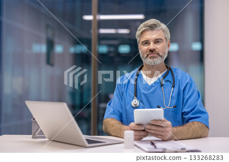 Experienced male doctor wearing scrubs and a stethoscope, looking at the camera while holding a digital tablet in a modern medical office, representing healthcare technology and digital medicine 133268283