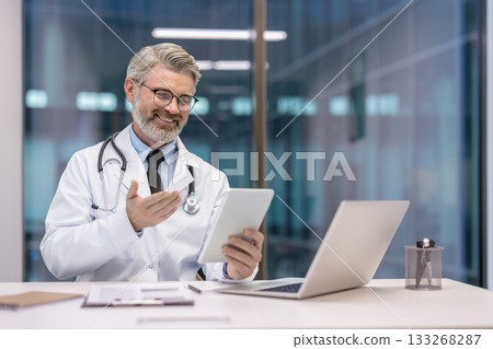 Male doctor in white coat and stethoscope smiles during a telemedicine video consultation, explaining treatment and offering remote care via tablet from a modern office setting Male doctor in white coat and stethoscope smiles during a telemedicine video consultation, explaining treatment and offering remote care via tablet from a modern office setting 133268287