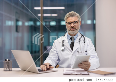 Professional senior male doctor wearing a white coat and stethoscope, sitting at a modern desk, working with a laptop and holding a digital tablet, providing healthcare and medical expertise 133268288