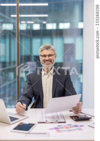 Mature businessman sitting at office desk, holding documents and pen, smiling at camera with a modern office interior background, representing professional success and corporate environment 133268298