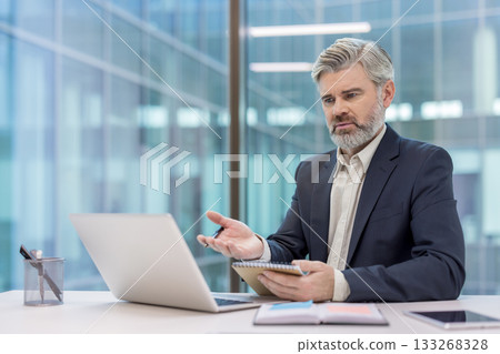 Mature businessman in suit on a remote video call from his office, gesturing with one hand while holding a notebook, focused and engaged in a virtual meeting discussion Mature businessman in suit on a remote video call from his office, gesturing with one hand while holding a notebook, focused and engaged in a virtual meeting discussion 133268328
