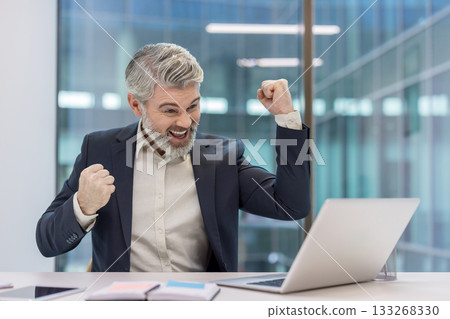 Excited mature businessman with a beard and gray hair smiling, raising fists in triumph while looking at a laptop screen in a modern office, celebrating business success or achievement 133268330