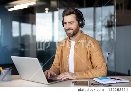 Happy bearded man in smart-casual attire wearing headphones, smiling at laptop during a virtual meeting or webinar in a bright modern office, focused and productive 133268334