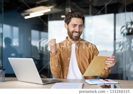 Happy adult man celebrating success, receiving good news by letter, reading a document at his office desk with a laptop and making a triumphant gesture Happy adult man celebrating success, receiving good news by letter, reading a document at his office desk with a laptop and making a triumphant gesture 133268335