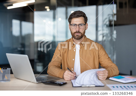 Businessman looking at camera, sitting at office desk, holding documents, writing on clipboard, with laptop and calculator in a modern office environment 133268346