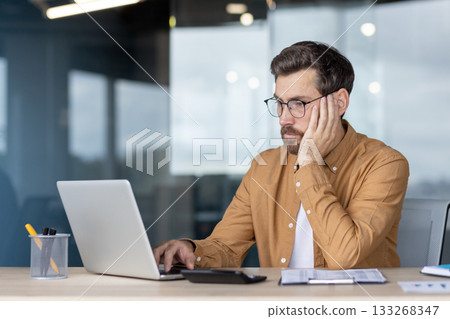 Businessman exhausted and bored at a desk, leaning on hand while typing on a laptop in a modern home office, showing stress, burnout and deadline pressure 133268347