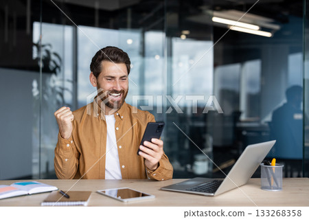 Smiling man feeling joy and success checking good news on smartphone, raising his fist in triumph while sitting at a desk in a modern office with a laptop Smiling man feeling joy and success checking good news on smartphone, raising his fist in triumph while sitting at a desk in a modern office with a laptop 133268358