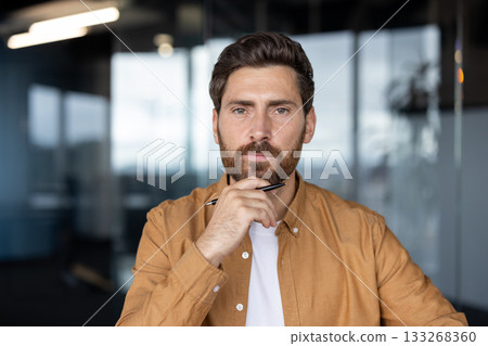 Serious well-groomed professional man having an idea, looking at camera while holding a pen under his chin, planning strategy in a contemporary workplace environment Serious well-groomed professional man having an idea, looking at camera while holding a pen under his chin, planning strategy in a contemporary workplace environment 133268360