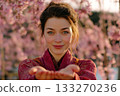 Smiling young woman in a red kimono holding out her hands with cherry blossoms in full bloom behind her 133270236