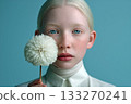 Minimalist portrait of an albino girl with light blonde hair and blue eyes holding a white flower against a turquoise background 133270241