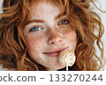 Closeup of a smiling young woman with red hair and freckles holding a white chocolate cake pop with sprinkles 133270244