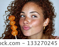 Playful young woman with freckles holding a skewer of candied fruit and making a pouty face against a plain background 133270245