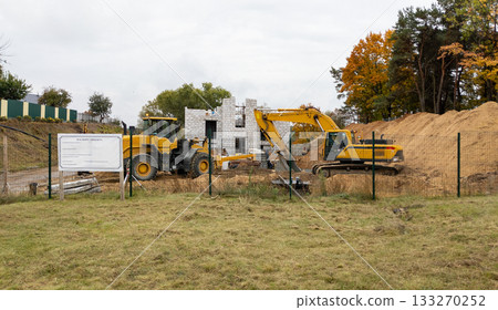Yellow excavator and loader against the backdrop of a brick building under construction and a pile of sand. Construction equipment Yellow excavator and loader against the backdrop of a brick building under construction and a pile of sand. Construction equipment 133270252