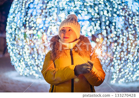 Young woman in knitted hat holds sparkler in her hands. Night festive street, shiny city decorations for Christmas. 133270428