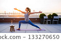 Woman practicing yoga warrior pose on rooftop at sunrise with city skyline image 133270650