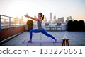 Woman practicing yoga warrior pose on rooftop with city skyline at sunset image 133270651