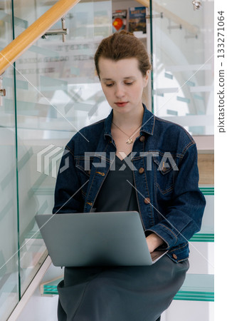 young woman is sitting on glass staircase with laptop in front of her and working.  133271064