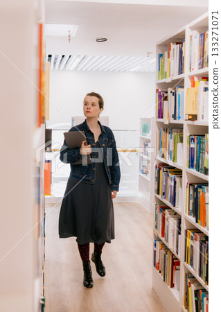 woman stands among shelves in library with laptop in hands, pointing book. university lecturer. 133271071