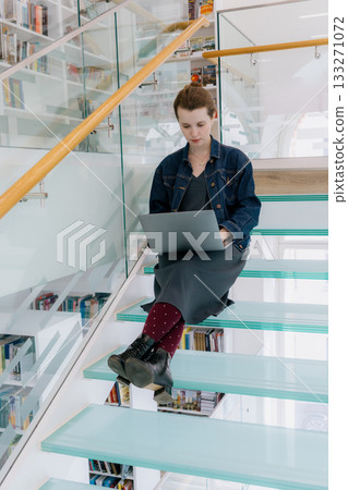 young woman is sitting on glass staircase with laptop in front of her and working.  133271072