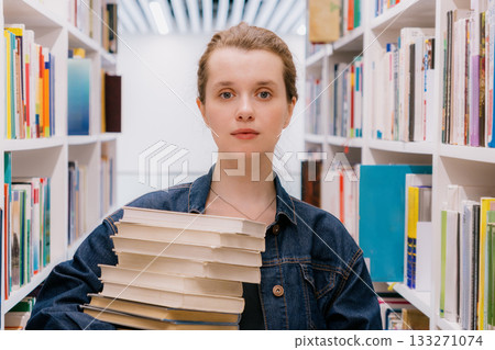 woman holds stack of books among shelves in library. student is getting education. 133271074