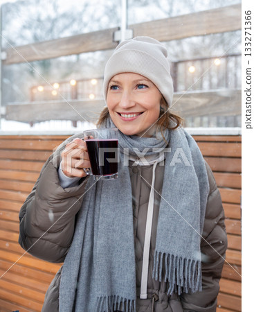 Snowy winter setting shows woman enjoying warm mulled wine with cozy smile. Soft lights, knit hat, scarf and puffer coat create festive Christmas atmosphere filled with comfort and gentle cold 133271366