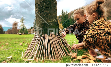 Children building a toy stick house around a tree in a park 133271367