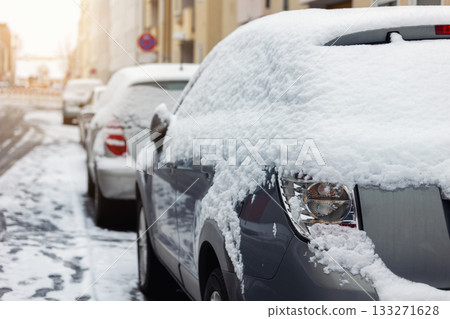 Urban street lined with parked cars covered in fresh snow under cold winter light. Silent morning atmosphere shows traces of traffic and frosty stillness between buildings and street lamps Urban street lined with parked cars covered in fresh snow under cold winter light. Silent morning atmosphere shows traces of traffic and frosty stillness between buildings and street lamps 133271628