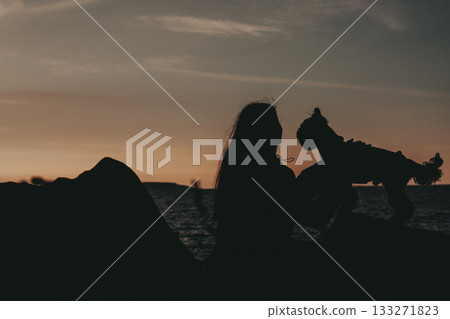 Silhouette of a woman and her small dog at sunset. A woman holds her Yorkshire terrier doggy up as they enjoy the sunset over the water 133271823