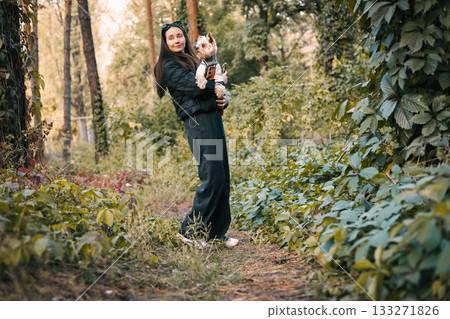 Young woman walking in the autumnal forest holding a Yorkshire Terrier dog. A stylish girl walking on a path in green forest with a lovable lapdog. 133271826