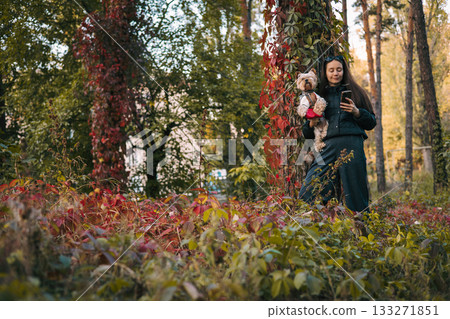 Young woman walking in autumn forest holding a Yorkshire Terrier dog and looking at smartphone. Relaxing walk with small dog in autumn woodland. 133271851
