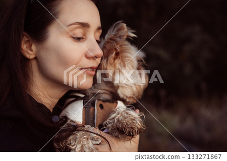 Woman lovingly embraces her small dog outdoors. A tender moment of affection. A girl is holding Yorkshire terrier doggy, enjoying a peaceful moment. Woman lovingly embraces her small dog outdoors. A tender moment of affection. A girl is holding Yorkshire terrier doggy, enjoying a peaceful moment. 133271867