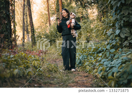 Young woman walking in the forest holding a Yorkshire Terrier dog and looking at smartphone. Outdoor lifestyle with pets, technology, and nature. Relaxing walk with small dog in autumn woodland. Young woman walking in the forest holding a Yorkshire Terrier dog and looking at smartphone. Outdoor lifestyle with pets, technology, and nature. Relaxing walk with small dog in autumn woodland. 133271875