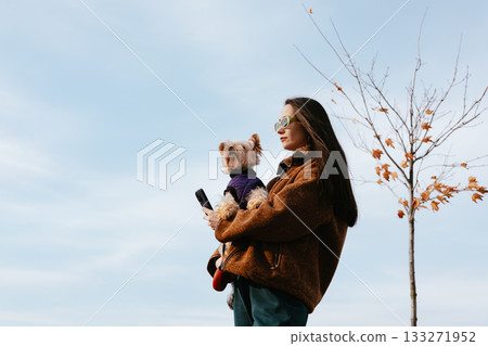 Young white woman enjoying autumn sunshine with a small dog outdoors, cozy fall outfit, happiness and slow living lifestyle moment, relaxation and mindfulness in nature. Pet and human companionship. 133271952