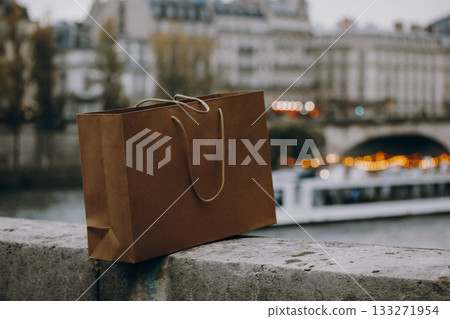 Brown eco-friendly paper shopping bag on a stone wall with blurred city lights and river in the background. Sustainable lifestyle, shopping, retail, and urban travel concept in Paris atmosphere. Brown eco-friendly paper shopping bag on a stone wall with blurred city lights and river in the background. Sustainable lifestyle, shopping, retail, and urban travel concept in Paris atmosphere. 133271954
