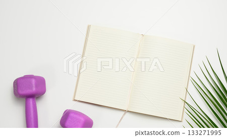 Open lined notebook with blank pages, surrounded by purple dumbbells and a green palm leaf on a white background. 133271995