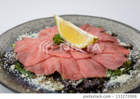 Tuna carpaccio with lemon and parmesan on greens, black plate, studio top view 133272008