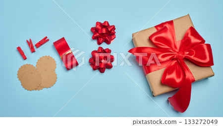 A gift box wrapped in kraft paper with a red satin bow, surrounded by packaging accessories such as tags, clothespins, and decorative bows. A gift box wrapped in kraft paper with a red satin bow, surrounded by packaging accessories such as tags, clothespins, and decorative bows. 133272049