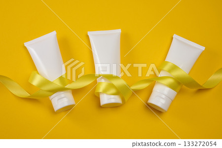 Three white plastic cosmetic tubes wrapped with a yellow satin ribbon on a yellow background. Gift, mockup, and personal care concept. 133272054