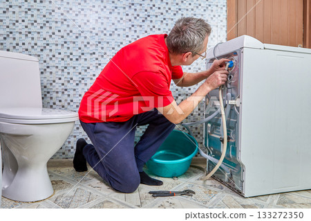 Plumber fastens water hoses to washing machine, hands connect fittings at rear panel, plumbing task involves water inlet hose installation. 133272350
