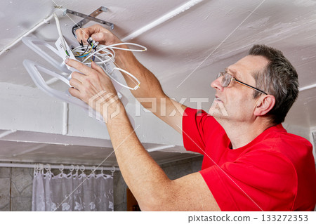 Worker installs LED chandelier on ceiling in countryside home, wires joined to contacts, mounting bracket secured, task nearing completion. 133272353
