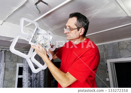 Fixing electrician holds new LED chandelier near ceiling in rural house, wires prepared for connection, fixture positioned for installation. Fixing electrician holds new LED chandelier near ceiling in rural house, wires prepared for connection, fixture positioned for installation. 133272358