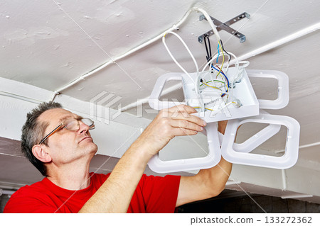 Positioning technician lifts LED chandelier toward ceiling in countryside home, wiring visible, structure aligned with mounting bracket. Positioning technician lifts LED chandelier toward ceiling in countryside home, wiring visible, structure aligned with mounting bracket. 133272362