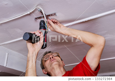 Tightening electrician uses cordless screwdriver to secure ceiling bracket for LED chandelier in rural house, cable group positioned for mounting. Tightening electrician uses cordless screwdriver to secure ceiling bracket for LED chandelier in rural house, cable group positioned for mounting. 133272364