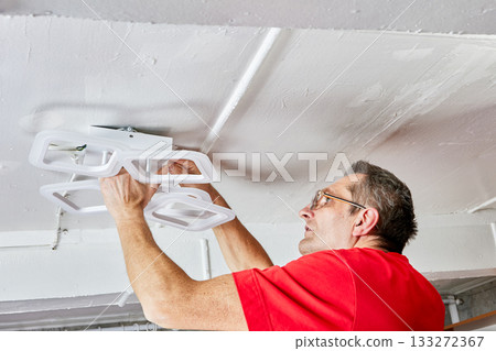 Installation process shows electrician mounting modern LED chandelier onto ceiling in rural house using both hands during connection of lighting fixture. Installation process shows electrician mounting modern LED chandelier onto ceiling in rural house using both hands during connection of lighting fixture. 133272367