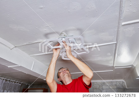 LED chandelier is positioned on ceiling while electrician performs installation in rural house connecting wires and securing fixture. 133272678
