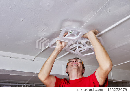 LED chandelier is set against ceiling during mounting work by electrician in rural house with hands reaching up for wiring. LED chandelier is set against ceiling during mounting work by electrician in rural house with hands reaching up for wiring. 133272679