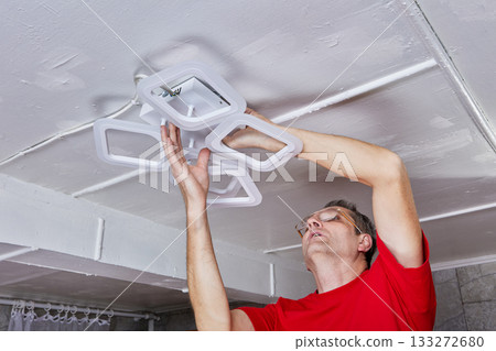 Fitting operation captures technician attaching new LED chandelier to ceiling in country home working with lighting unit during ceiling installation phase. 133272680